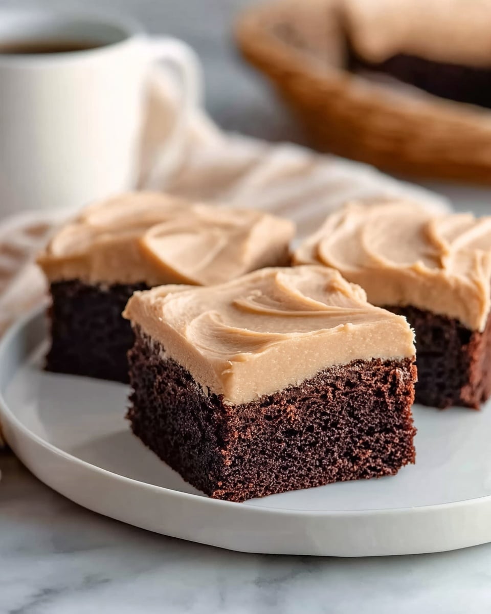 Three square pieces of chocolate cake with one thick layer of light brown frosting on top, sitting close together on a white plate. The cake layers are dark brown with a soft, moist texture, and the frosting is smooth with slight swirls and a creamy look. The plate is placed on a white marbled surface with a soft blur in the background, showing hints of a white cup and a basket. Photo taken with an iphone --ar 4:5 --v 7
