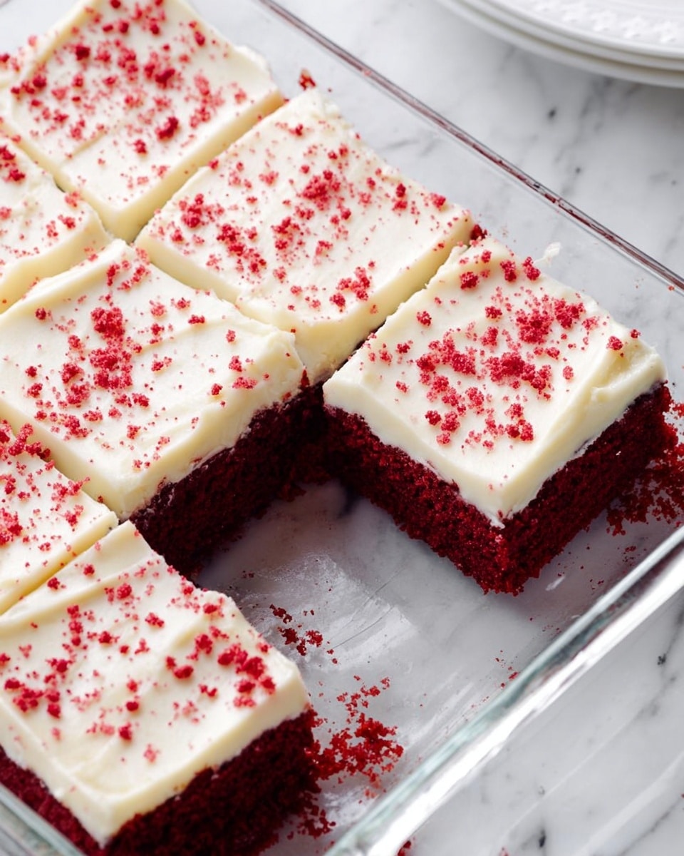 A clear glass baking dish contains a red velvet cake cut into nine square pieces, with one piece slightly separated from the rest. The cake has two visible layers: a thick, dark red base with a soft, crumbly texture, and a smooth, creamy white frosting layer on top. Small red sprinkles are scattered evenly across the frosting, adding a delicate texture. The dish is placed on a white marbled surface, and a small part of a white plate is visible in the top right corner. Photo taken with an iphone --ar 4:5 --v 7