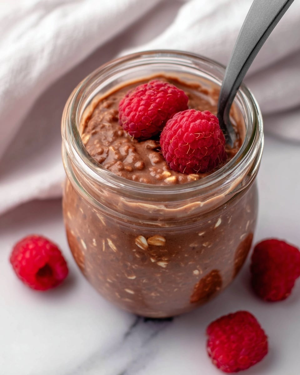 The image shows a clear glass jar filled with a thick, brown mixture that looks like chocolate oatmeal, with visible oats mixed inside. On top of the mixture are three bright red raspberries, adding a pop of color. A dark gray spoon is dipped into the jar from the right side, partially covered by the oatmeal. Around the jar, there are a few more raspberries placed on a white marbled surface with a soft white cloth in the background. The photo is close-up and focused on the top and side of the jar. photo taken with an iphone --ar 4:5 --v 7