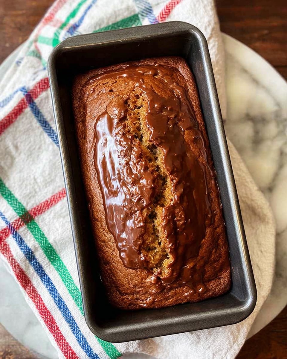 A close-up view of a single loaf of chocolate-glazed banana bread resting inside a dark metal loaf pan. The bread has a golden-brown top with a moist texture visible from cracks and a shiny chocolate glaze that is thickly spread, pooling slightly around the edges inside the pan. The pan sits on a white marbled surface partially covered by a soft white cloth with red, green, and blue stripe patterns. photo taken with an iphone --ar 4:5 --v 7