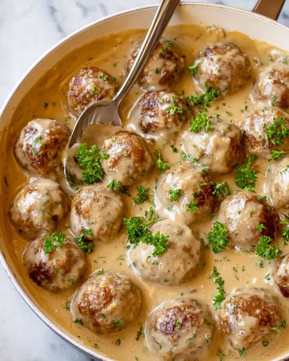 A white pan filled with many round meatballs covered in a smooth, creamy light brown sauce, speckled with small green herb pieces. The meatballs are mostly submerged, but their browned tops show through the thick sauce, which has a slightly glossy texture. Bright green chopped parsley is sprinkled on top, adding a fresh contrast. A silver spoon rests on the side of the pan. The scene is set on a white marbled surface. photo taken with an iphone --ar 4:5 --v 7