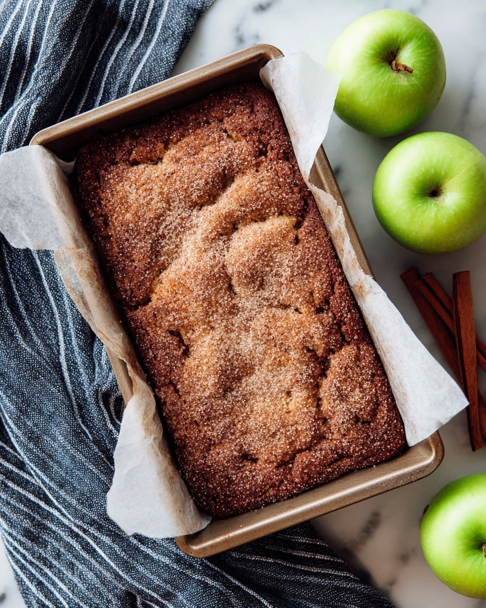 A rectangular brown baked apple cinnamon cake sits inside a baking pan lined with white parchment paper, with a rough and slightly cracked top texture covered in a layer of granulated sugar and cinnamon giving a sparkling look. The pan rests on a white marbled surface next to two bright green apples and two brown cinnamon sticks. A navy blue and white striped cloth is partly draped around the lower left corner of the pan. photo taken with an iphone --ar 4:5 --v 7