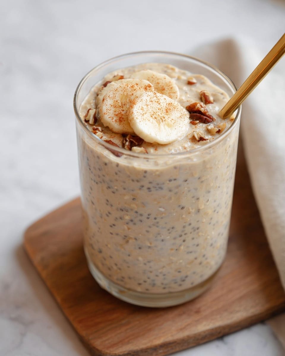 A tall clear glass filled with creamy beige overnight oats mixed with small dark chia seeds shows a thick, smooth texture with some chunks. On top, three thin banana slices are neatly placed in a small stack, lightly dusted with a sprinkle of reddish cinnamon powder and small chopped nuts. A shiny gold spoon rests inside the glass, leaning against the rim on the right side. The glass is set on a small wooden board, with a soft white marbled surface in the background. photo taken with an iphone --ar 4:5 --v 7