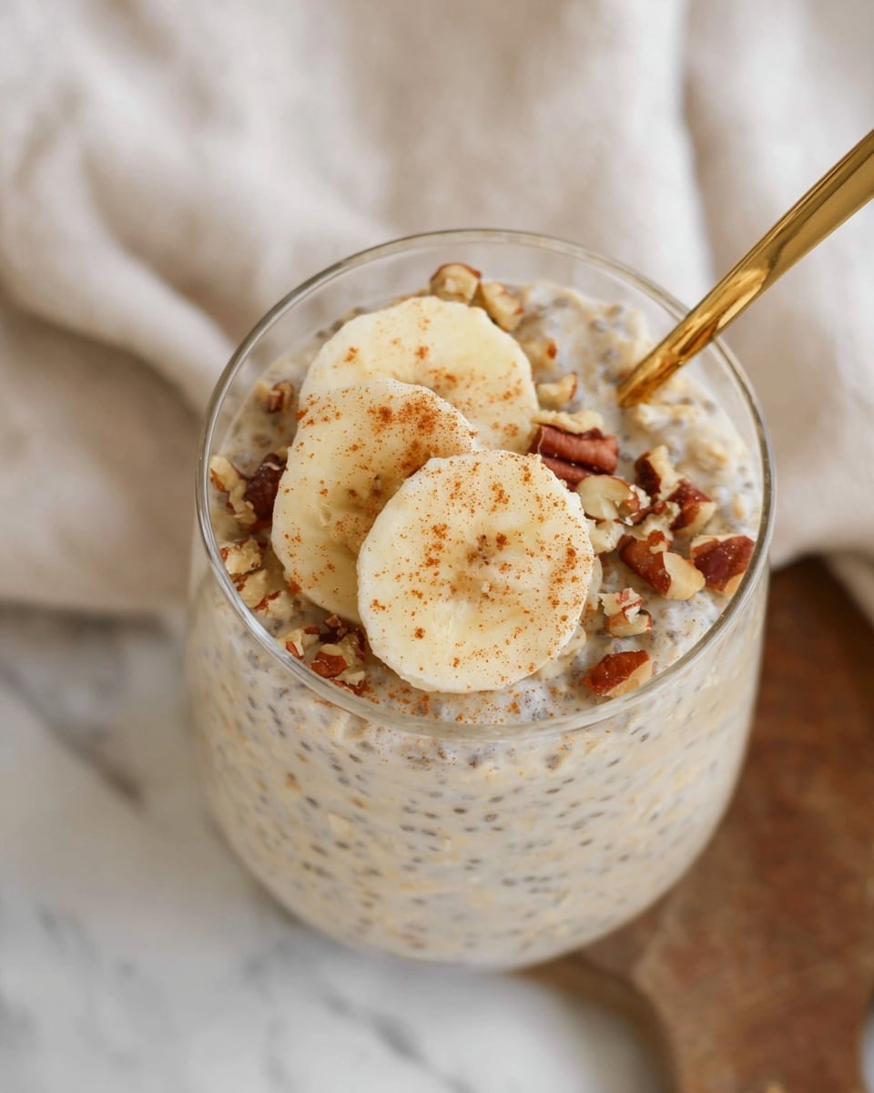A clear glass filled with creamy oatmeal mixed with visible chia seeds and nuts, creating a speckled, slightly chunky texture. On top, there are three round slices of banana neatly placed in the center, sprinkled with small pieces of chopped nuts and a light dusting of cinnamon. A gold spoon lies partially submerged on the right side of the glass. The background is a white marbled texture with a soft beige cloth and wooden board softly blurred in the back. photo taken with an iphone --ar 4:5 --v 7