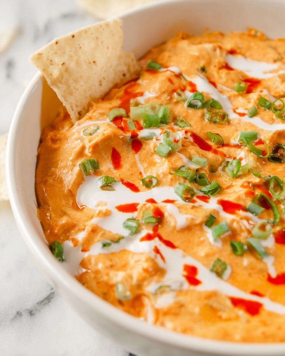 The dish is a white oval baking dish filled with a thick, creamy orange dip, likely made of cheese and buffalo sauce, visible from its slightly textured surface. The top layer is decorated with thin white sauce drizzles unevenly spread across the dip and scattered small green onion pieces adding a fresh contrast. There are also several streaks of red hot sauce on top. The dish is placed on a white marbled surface, accompanied by off-white tortilla chips scattered beside it, a small white bowl with bright orange baby carrots, and another white bowl with fresh cucumber slices. photo taken with an iphone --ar 4:5 --v 7