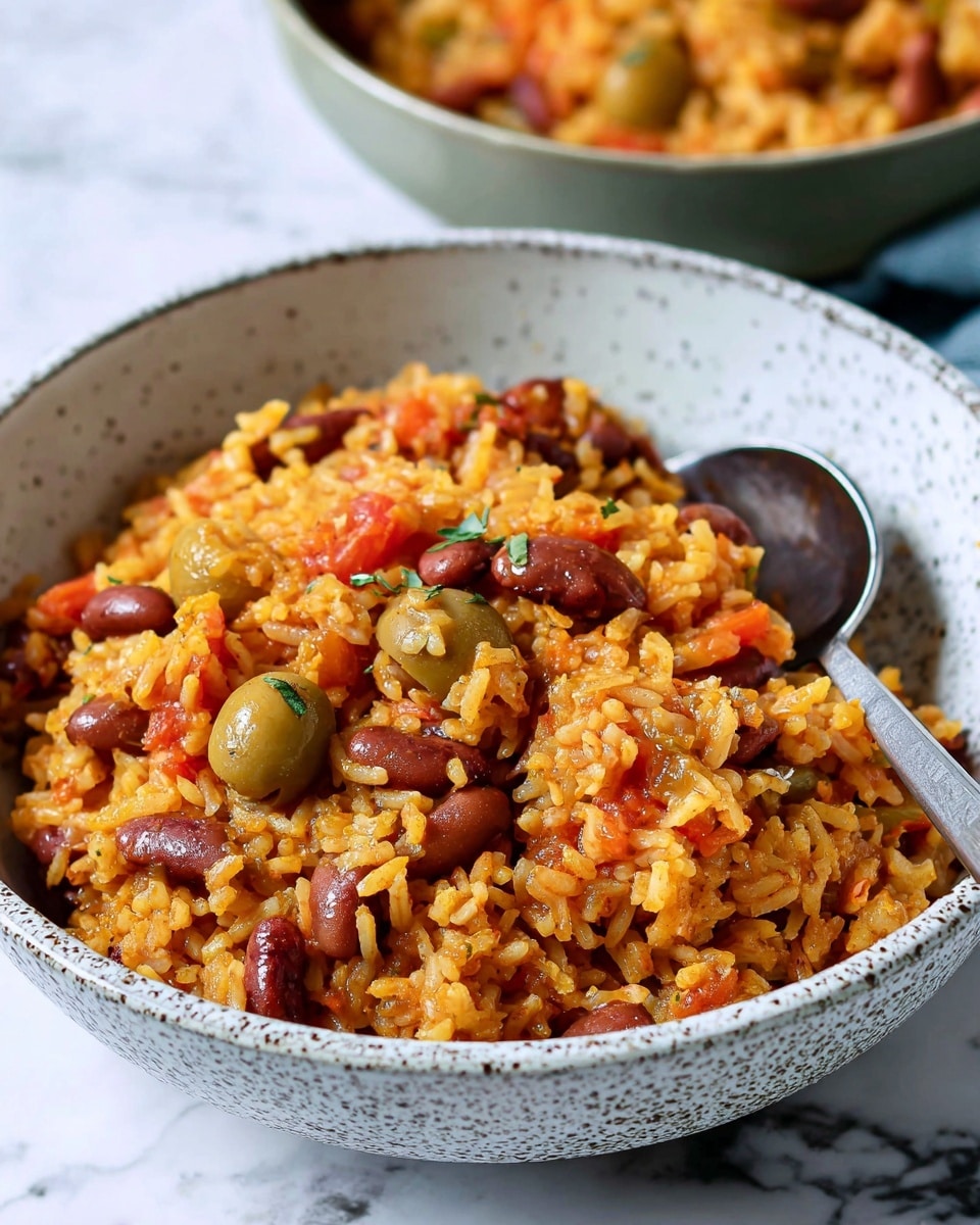 A close-up of a dish in a white bowl with a speckled texture, filled with a mix of yellow-orange cooked rice and red beans. The rice has small pieces of cooked red tomatoes and green olives scattered throughout, adding pops of color and texture. A silver spoon is partially immersed in the rice and beans, resting inside the bowl. The background shows a blurred second bowl with the same dish and a white marbled surface underneath. Photo taken with an iphone --ar 4:5 --v 7