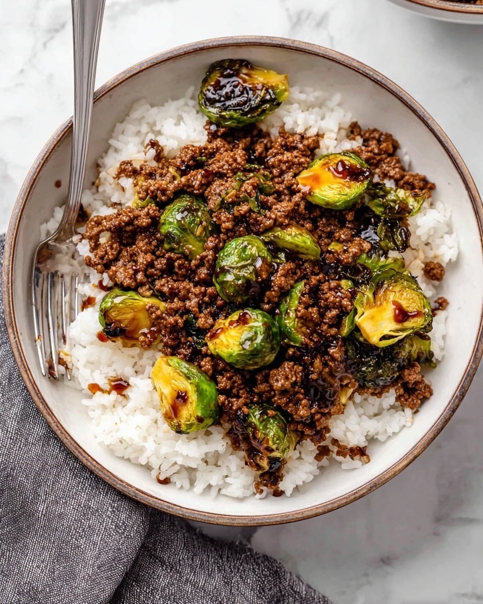 A white bowl with a brown rim filled with a base layer of fluffy white rice, topped with cooked ground beef that is brown and crumbly, mixed with bright green Brussels sprouts that are slightly charred and yellowed on some leaves, and drizzled with a dark glossy sauce. A silver fork rests in the bowl on the left side. The bowl is placed on a white marbled surface with a gray cloth napkin beside it. photo taken with an iphone --ar 4:5 --v 7