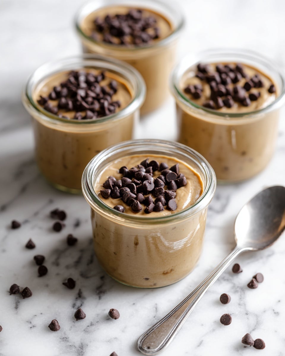 Three clear glass jars filled with creamy light brown pudding sit on a white marbled surface. Each jar has a smooth pudding layer topped generously with small, dark chocolate chips. Scattered chocolate chips surround the jars on the surface. A silver spoon lies next to the jar in the front right, its shiny texture contrasting with the soft pudding. The jars are arranged close to each other, showing details of the pudding’s soft texture inside the glass. Photo taken with an iphone --ar 4:5 --v 7