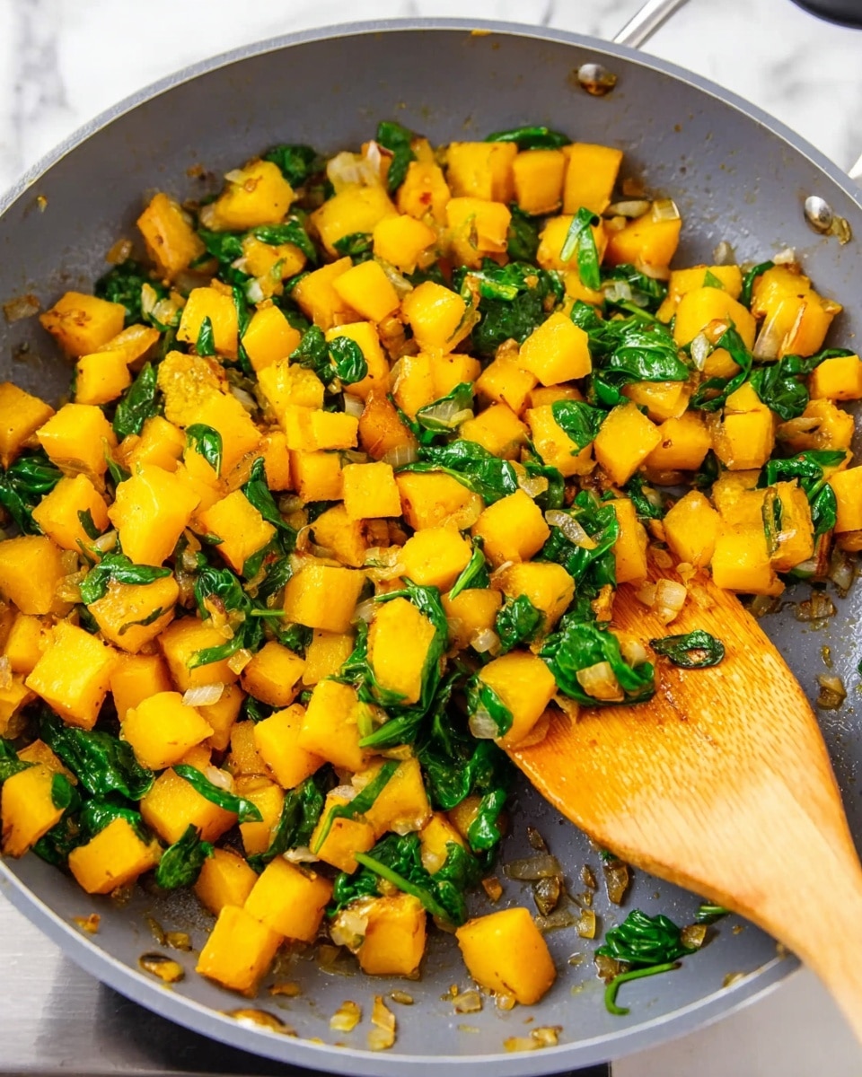 A close-up view of a frying pan filled with diced golden yellow squash pieces mixed with bright green spinach leaves and some small bits of cooked onions scattered around, all showing a warm, slightly sautéed texture. A wooden spatula with a smooth, slightly stained surface rests inside the pan on the right side, partially lifted over the veggies. The pan itself is gray with a black handle, and the background shows a white marbled texture counter top. Photo taken with an iphone --ar 4:5 --v 7
