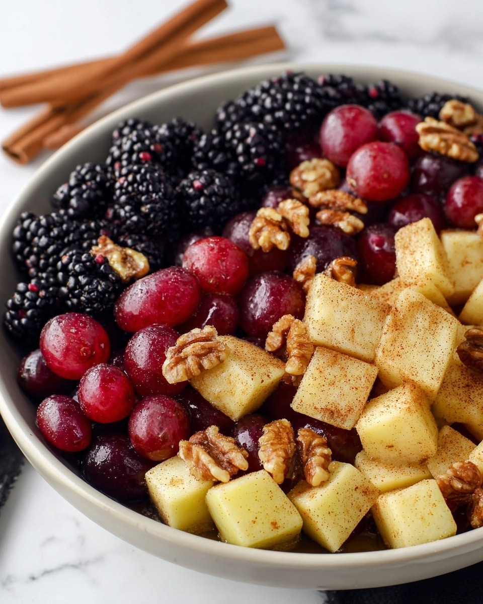 A close-up view of a white bowl filled with a colorful fruit salad that includes three main layers: the first layer consists of small blackberries with a bumpy texture and deep black-purple color; the second layer is made of red grapes, smooth and shiny with a rich red hue; and the third layer has pale yellow apple cubes, coated with a light dusting of cinnamon giving them a speckled brown look. Scattered throughout the fruit are walnut halves that add a rough texture and warm brown color, complementing the fruity layers. The bowl sits on a white marbled surface and there is a pair of cinnamon sticks placed nearby. Photo taken with an iphone --ar 4:5 --v 7