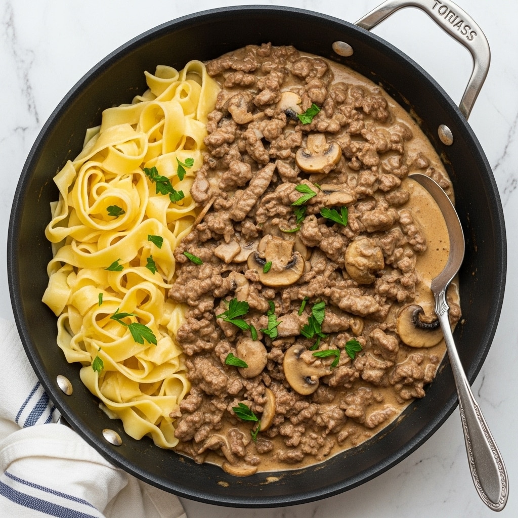 A black skillet filled with wide, pale yellow pasta ribbons folded on the left side, covered by a thick, creamy light brown sauce with many pieces of browned beef and sliced mushrooms mixed in, sprinkled with small bright green parsley leaves on top; a silver spoon rests on the right side inside the skillet, all placed on a white marbled surface with a white and blue striped cloth nearby, photo taken with an iphone --ar 4:5 --v 7
