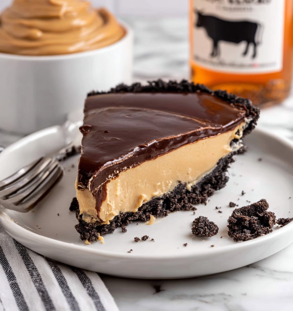 A slice of pie sits on a white plate with an uneven dark chocolate crumb crust at the bottom, topped by a thick, smooth layer of creamy light caramel-colored filling in the middle. Above the filling, a glossy dark chocolate ganache layer covers the top, with slight ripples on its surface. Around the plate, a few small chocolate crumbs are scattered. In the background on a white marbled surface, there is a white bowl filled with swirled light caramel creamy spread and a bottle with an orange liquid and a white label featuring a black and white cow illustration. A silver fork rests on the left side of the plate, and a striped white and black cloth can be seen at the bottom left corner. Photo taken with an iphone --ar 4:5 --v 7