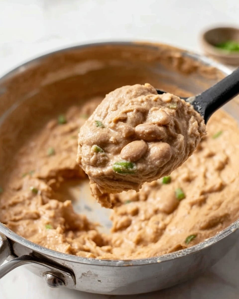 The image shows a close-up of creamy refried beans in a silver metal pan with a black plastic spoon lifting a scoop of the beans. The refried beans are light brown, thick, and have a smooth texture with some small bean pieces. There are tiny green bits mixed in, likely chopped herbs or peppers. The background surface is a white marbled texture, and the pan is placed on top of it. photo taken with an iphone --ar 4:5 --v 7