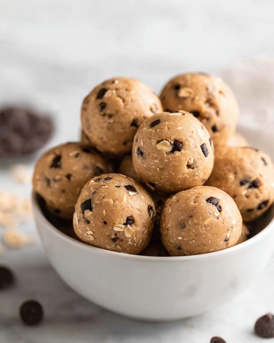 A white bowl filled with several round cookie dough balls stacked closely together, each ball showing a light brown color with visible small dark chocolate chips scattered throughout. The texture of the dough appears soft and slightly smooth with small oats or grain pieces mixed in. The bowl sits on a white marbled surface, and the background is softly blurred with hints of dark chocolate chips. photo taken with an iphone --ar 4:5 --v 7