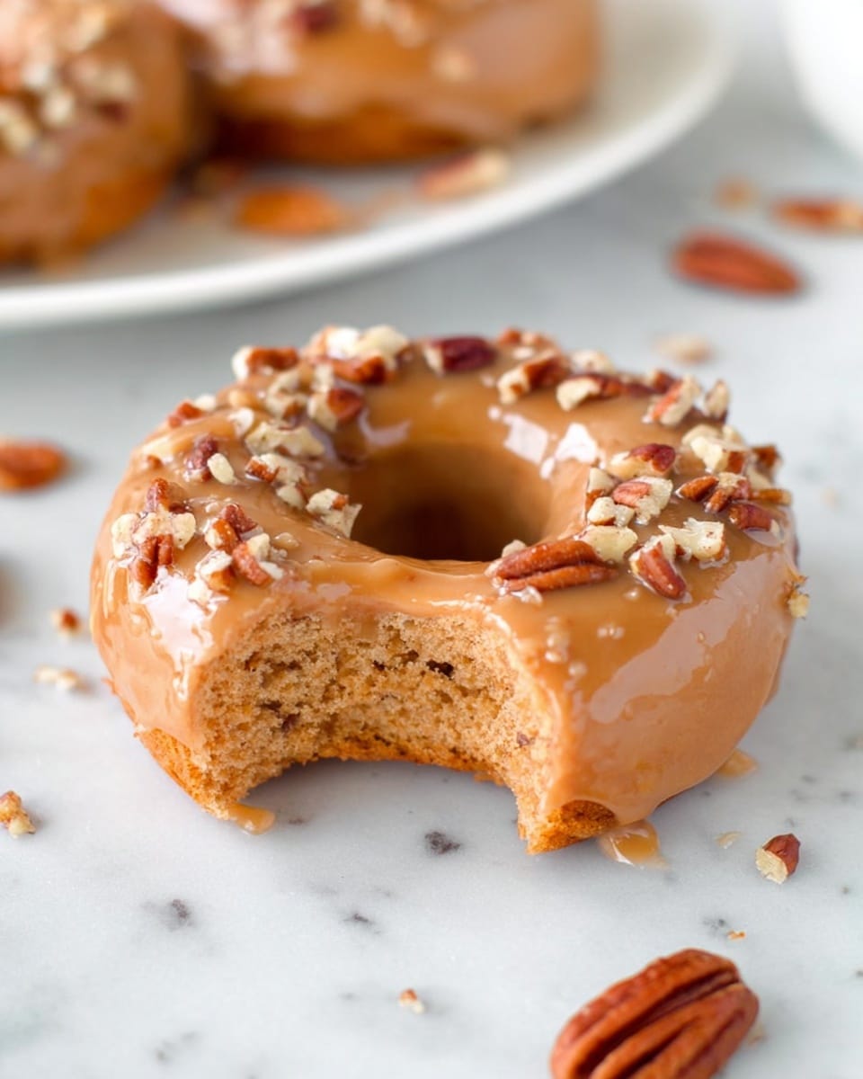 A single donut with one bite taken out sits on a white marbled surface. The donut has a light brown color with a smooth, shiny caramel glaze covering its top layer. On the glaze, there are small pieces of chopped pecans sprinkled around the upper edge. The inside of the donut shows a soft, fluffy texture with a golden brown hue. There are a few scattered whole pecans in the background on the white marbled surface. photo taken with an iphone --ar 4:5 --v 7