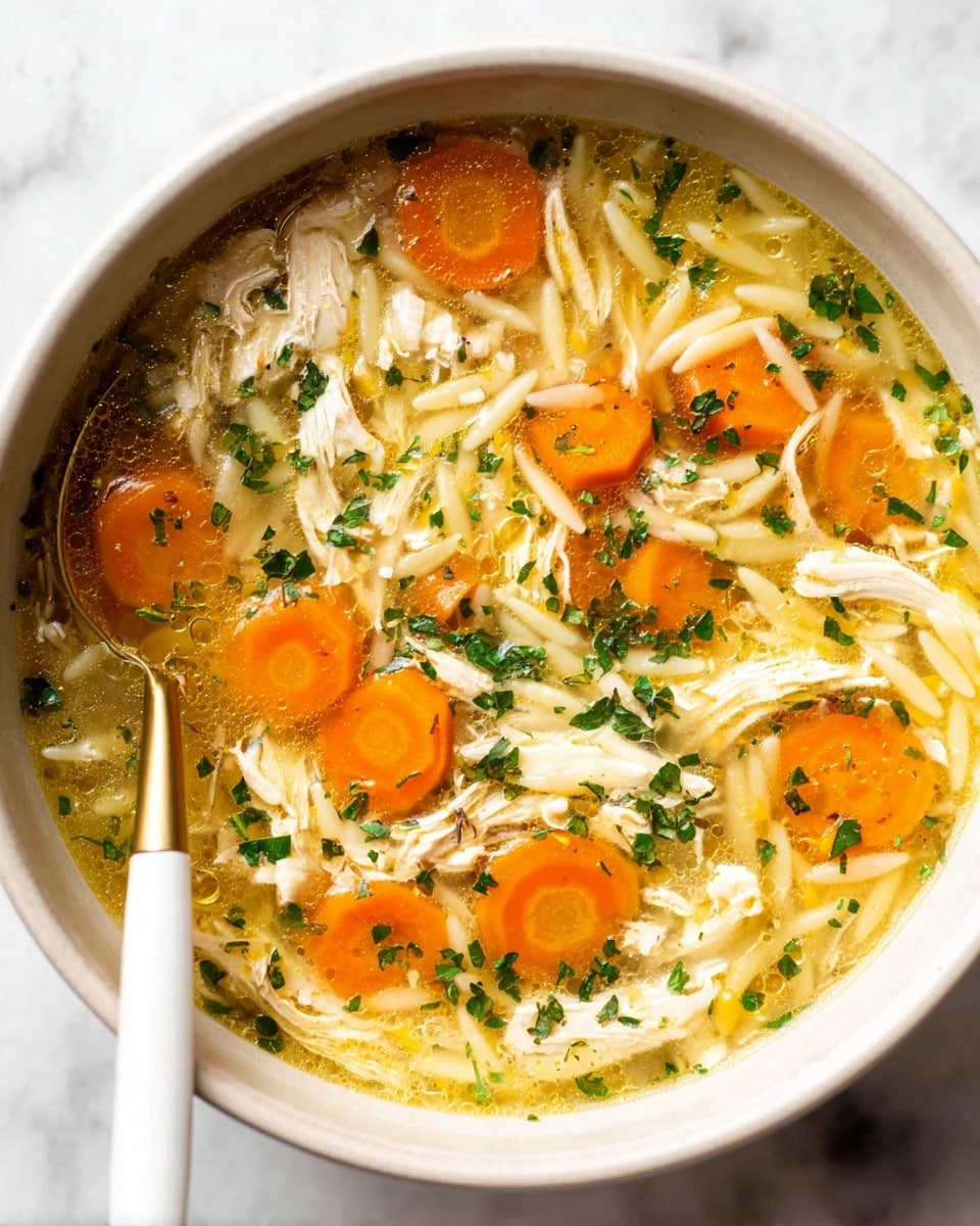 A close-up view of a white bowl filled with clear chicken broth soup showing multiple layers of ingredients; the bottom layer is the light golden clear broth, floating on top are bright orange round carrot slices evenly spread, soft white shredded chicken pieces scattered across, and small pale yellow orzo pasta pieces mixed in; finely chopped green herbs are sprinkled over the entire surface adding texture and color contrast. A golden spoon with a white handle rests inside the bowl on the left side, partially submerged in the soup. The bowl sits on a white marbled surface. photo taken with an iphone --ar 4:5 --v 7