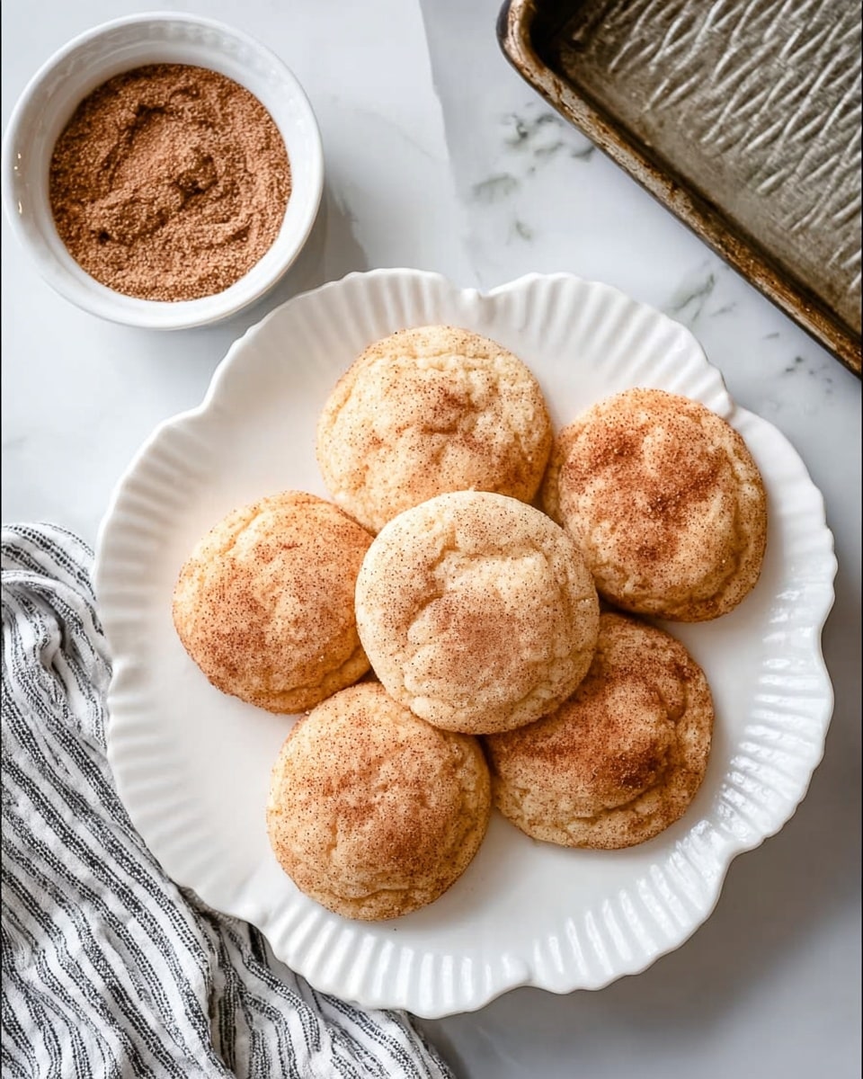 A stack of five round snickerdoodle cookies sits centered on a white plate with a raised floral pattern. Each cookie has a pale golden color and is dusted evenly with a light cinnamon sugar coating, giving them a slightly textured surface. The cookies are soft and slightly cracked on top, stacked perfectly with edges showing a gentle, uneven thickness. The plate and cookies rest on a white marbled surface that adds a clean, elegant look. Photo taken with an iphone --ar 4:5 --v 7