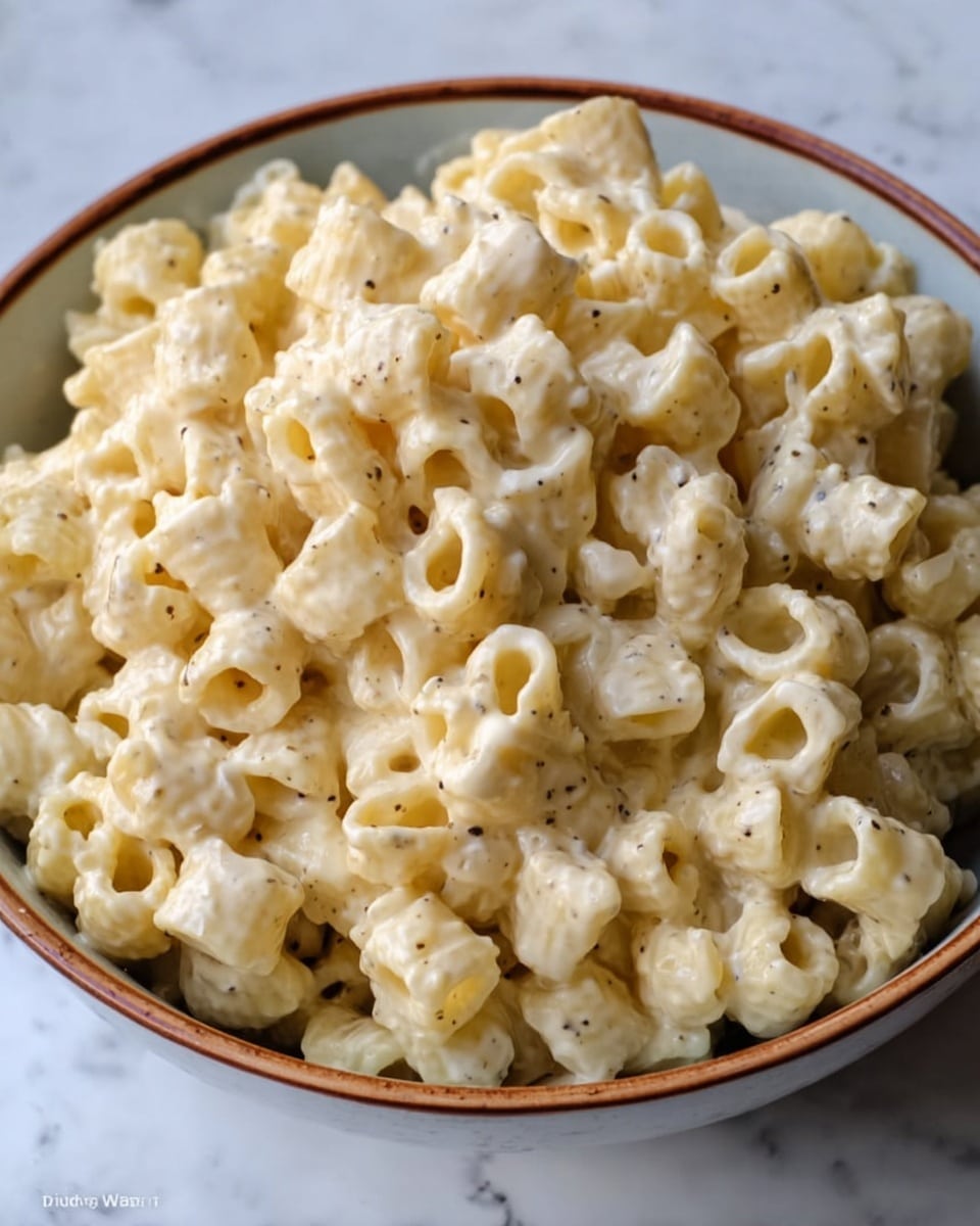 The image shows a white ceramic bowl filled with small tube-shaped pasta covered in a thick creamy sauce. The pasta is pale yellow with a smooth, slightly glossy texture, and the sauce is off-white with small specks of black pepper spread throughout. The bowl has a clean, simple design with a thin brown rim. The pasta fills the bowl fully, piled high and looks soft and rich. The background is a white marbled texture, giving a clean and bright look. photo taken with an iphone --ar 4:5 --v 7
