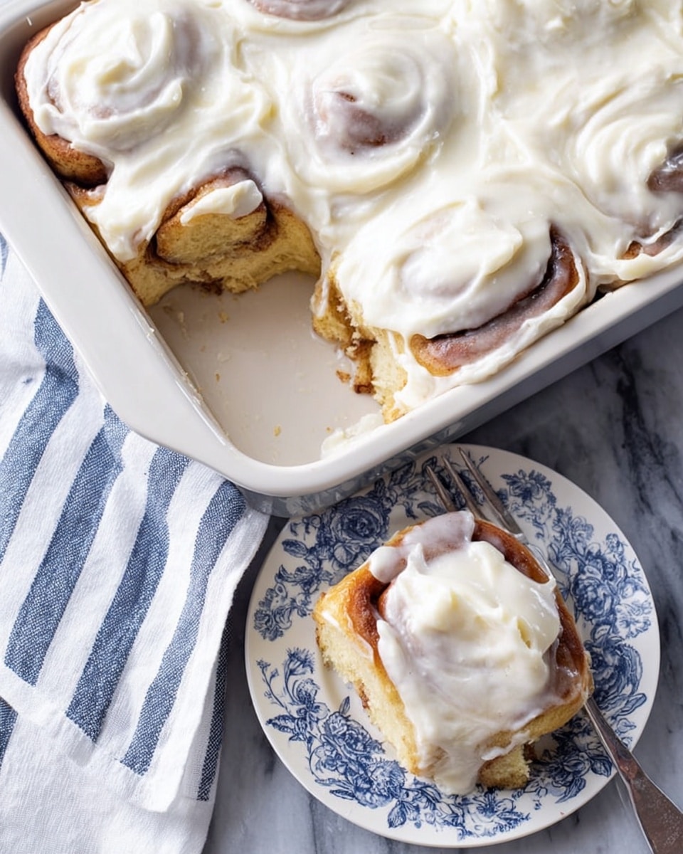 A square white baking dish holds several soft cinnamon rolls covered thickly with smooth white cream cheese frosting, which is spread unevenly showing parts of the golden brown rolls below. One cinnamon roll is cut out, revealing a sticky, gooey inner swirl with a light brown color and topped with the same thick frosting. This single roll sits on a small white plate decorated with blue floral patterns, placed on a white marbled textured surface next to a white cloth with blue stripes. photo taken with an iphone --ar 4:5 --v 7