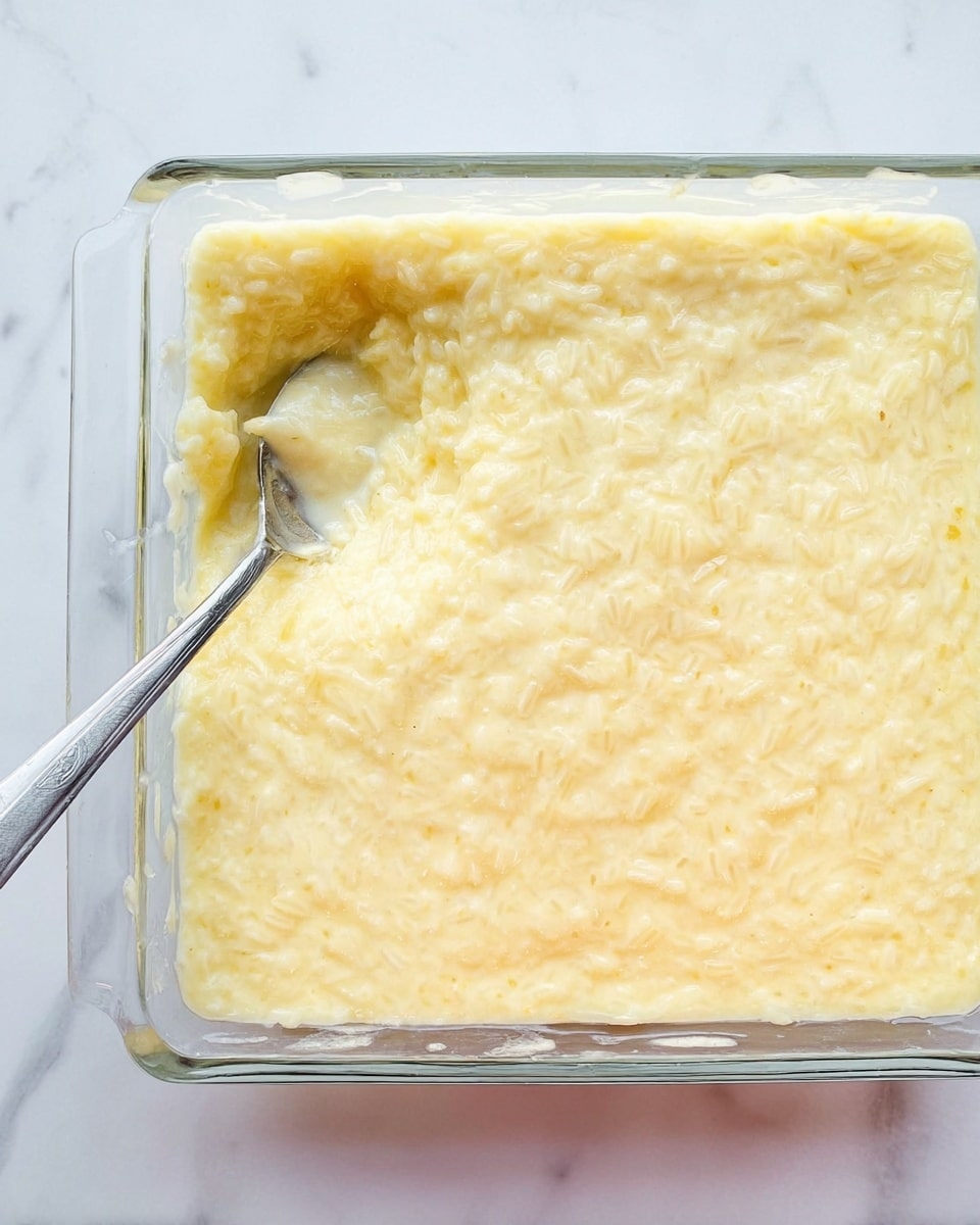 A clear square glass baking dish filled with creamy rice pudding, showing one thick, smooth, pale yellow layer with visible rice grains evenly spread across the top. A silver spoon is partially submerged on the left side, with a small scoop taken out. The dish rests on a white marbled surface. Photo taken with an iphone --ar 4:5 --v 7