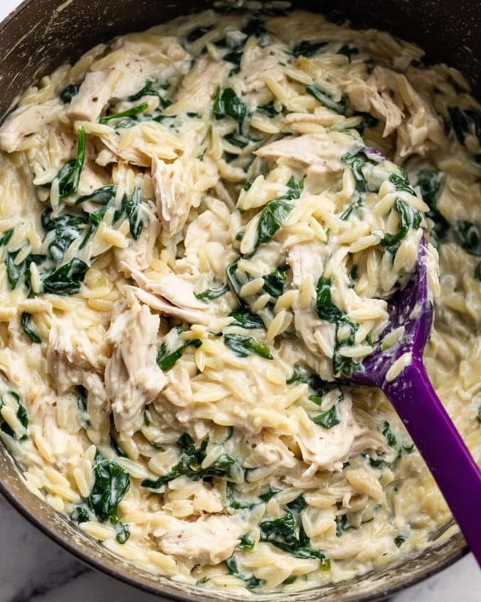 The image shows a close-up of a creamy pasta dish in a pot. The dish has two main layers: a creamy white sauce coating the orzo pasta, which looks soft and smooth, and green leafy spinach mixed evenly throughout, adding contrast. There are also pieces of white shredded chicken scattered on top, giving texture and variation. A purple spoon is partially visible on the right side, scooping the dish. The background surface is white marble. Photo taken with an iphone --ar 4:5 --v 7