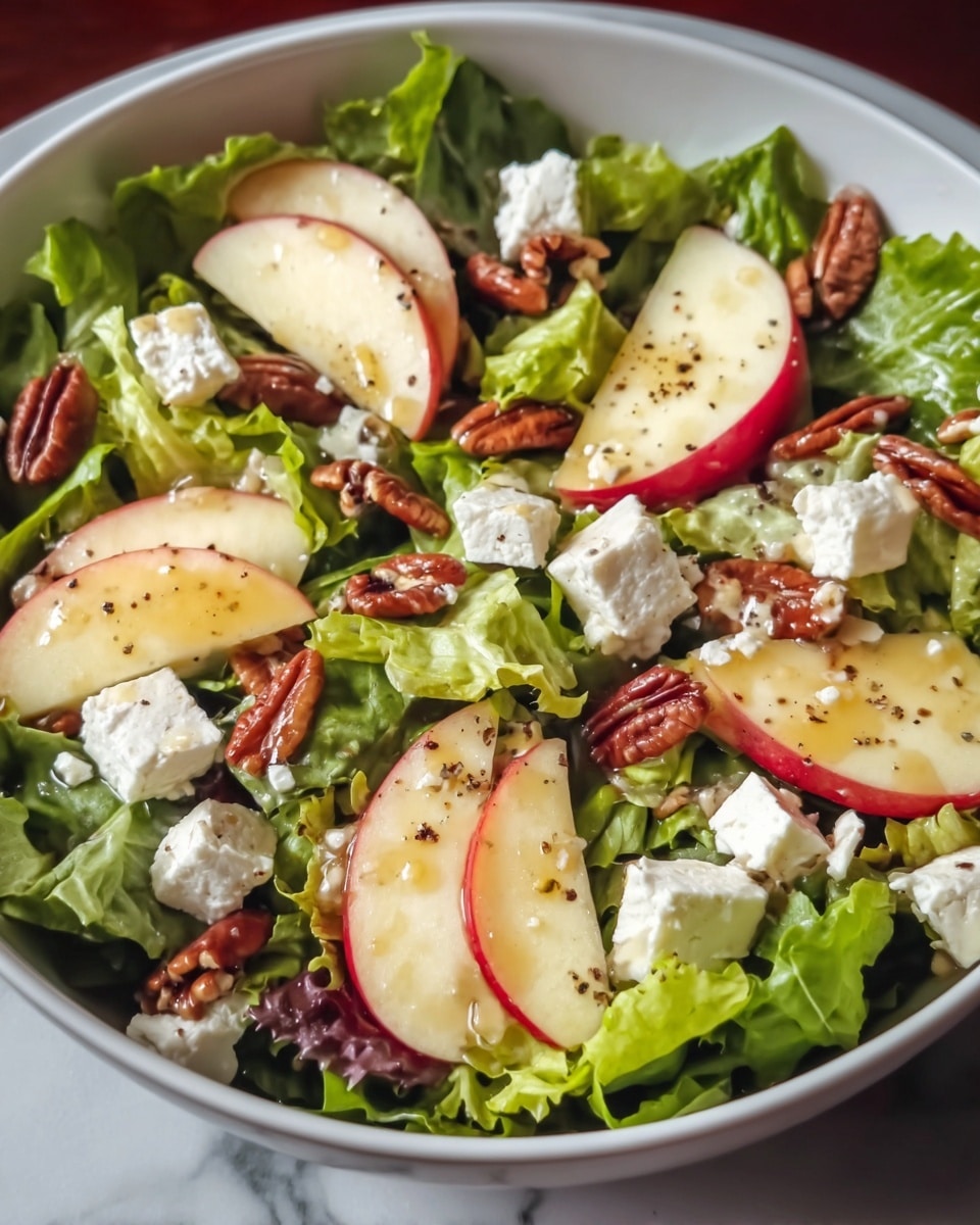 A white bowl is filled with fresh green leafy lettuce as the base layer, topped with evenly spaced slices of red-skinned apple showing their pale cream insides. Scattered over the apples and lettuce are small clusters of white feta cheese cubes and roughly chopped brown pecan nuts. The salad is lightly sprinkled with black pepper and a shiny drizzle of dressing that adds a slight gloss to some parts. The bowl sits on a white marbled surface. photo taken with an iphone --ar 4:5 --v 7