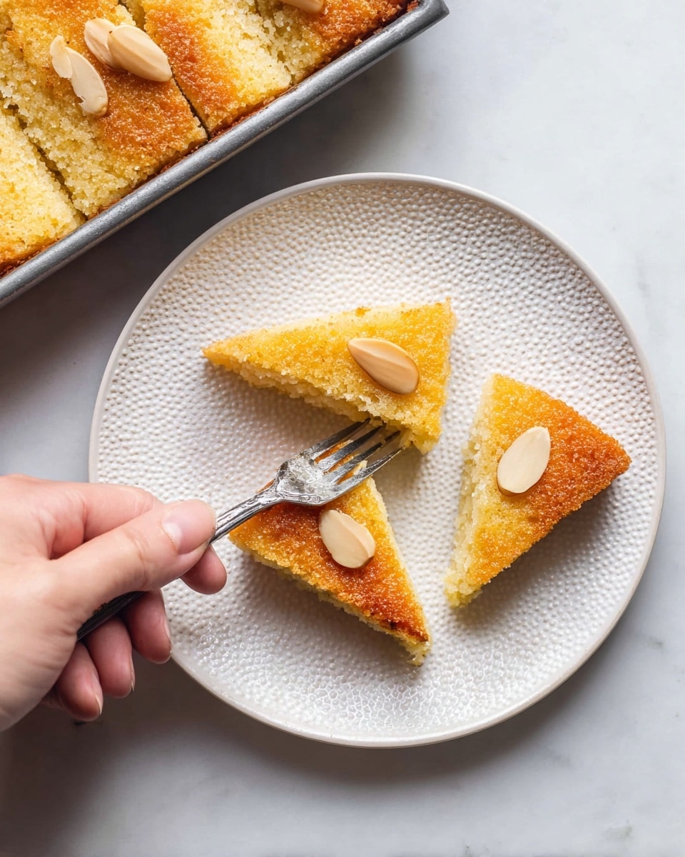 Three diamond-shaped pieces of golden brown cake with a slightly coarse texture sit on a white textured plate. Each piece has a single blanched almond placed in the center. A woman's hand holds a fork, cutting into one of the pieces, revealing a light, soft interior. In the top left corner of the image, part of a metal pan with more pieces of the same cake is visible. The whole setting is on a white marbled surface. photo taken with an iphone --ar 4:5 --v 7