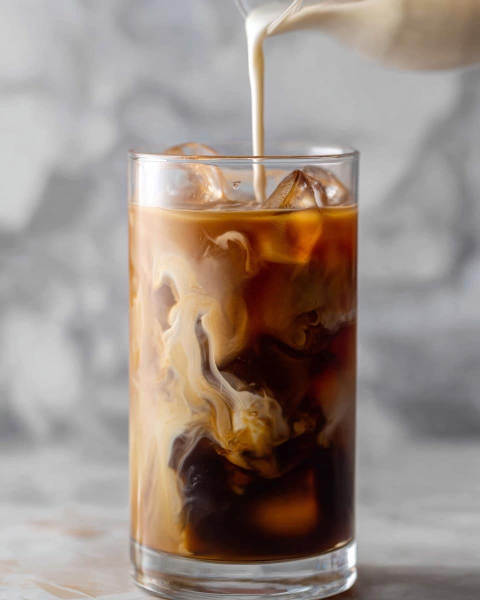 A tall clear glass filled with dark black coffee and clear ice cubes inside, with a stream of creamy white milk pouring into the coffee from above, creating swirling patterns of light brown and cream that mix with the dark liquid. The glass sits on a white marbled surface scattered with a few coffee beans, and the background shows soft warm blurred lights. photo taken with an iphone --ar 4:5 --v 7