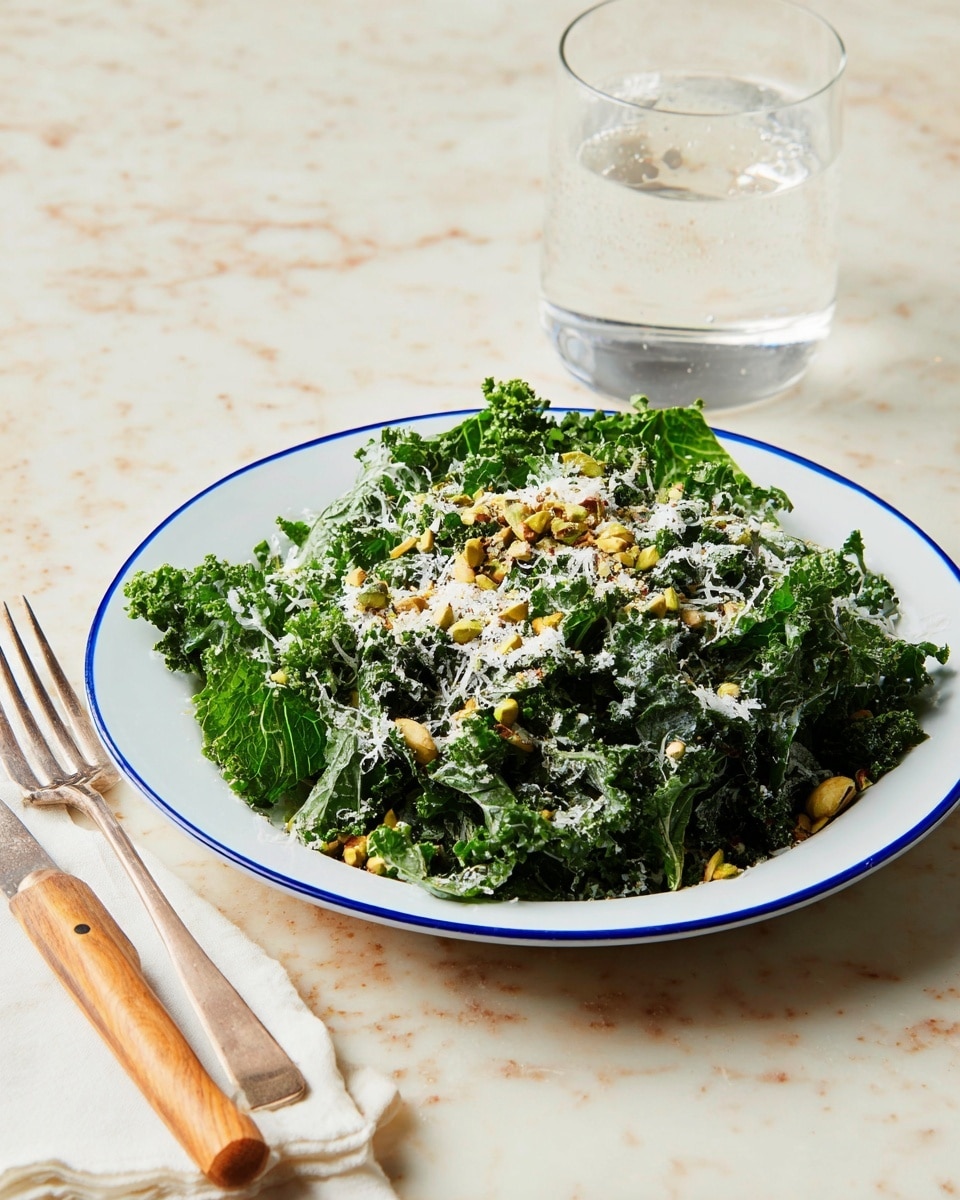 A white plate with a blue rim holds a fresh kale salad made of dark green, curly kale leaves as the first layer. On top, there is a sprinkling of light green chopped pistachios scattered unevenly. Over the nuts, there is a fine dusting of white grated cheese covering the surface of the salad. The plate sits on a white marbled textured surface, next to a fork with a wooden handle and a clear glass of sparkling water filled halfway, all lit by soft natural light. Photo taken with an iphone --ar 4:5 --v 7