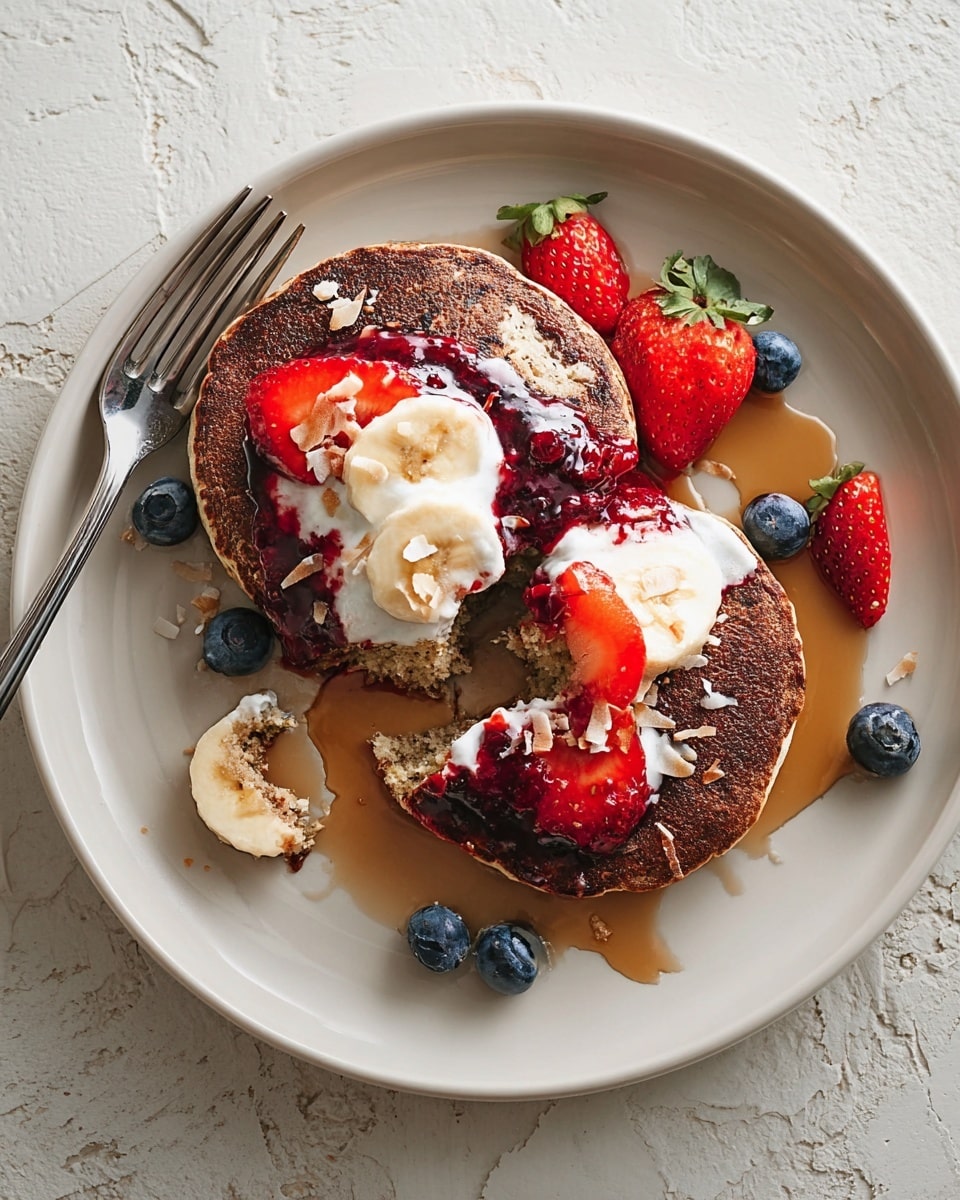 Two round, dark brown pancakes sit on a white plate with a white marbled texture underneath. The top pancake is plain with a few scattered toasted coconut flakes. The bottom pancake is partly covered with a layer of white cream, a thick red berry sauce, and fresh blueberries. There are strawberry and banana slices placed on and around the pancakes, along with more blueberries scattered on the plate. Light brown syrup pools on the plate, partly soaking the pancakes. A silver fork lies near a small broken piece of pancake with cream on it. Photo taken with an iphone --ar 4:5 --v 7